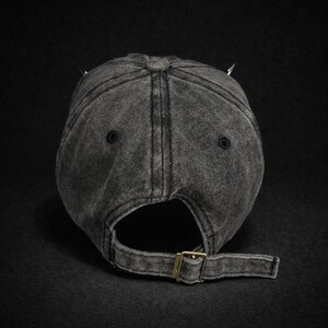 May include: A dark gray, distressed baseball cap with a curved brim and adjustable strap. The cap features two silver-colored spikes on top. The back of the cap has a metal buckle for adjustment.