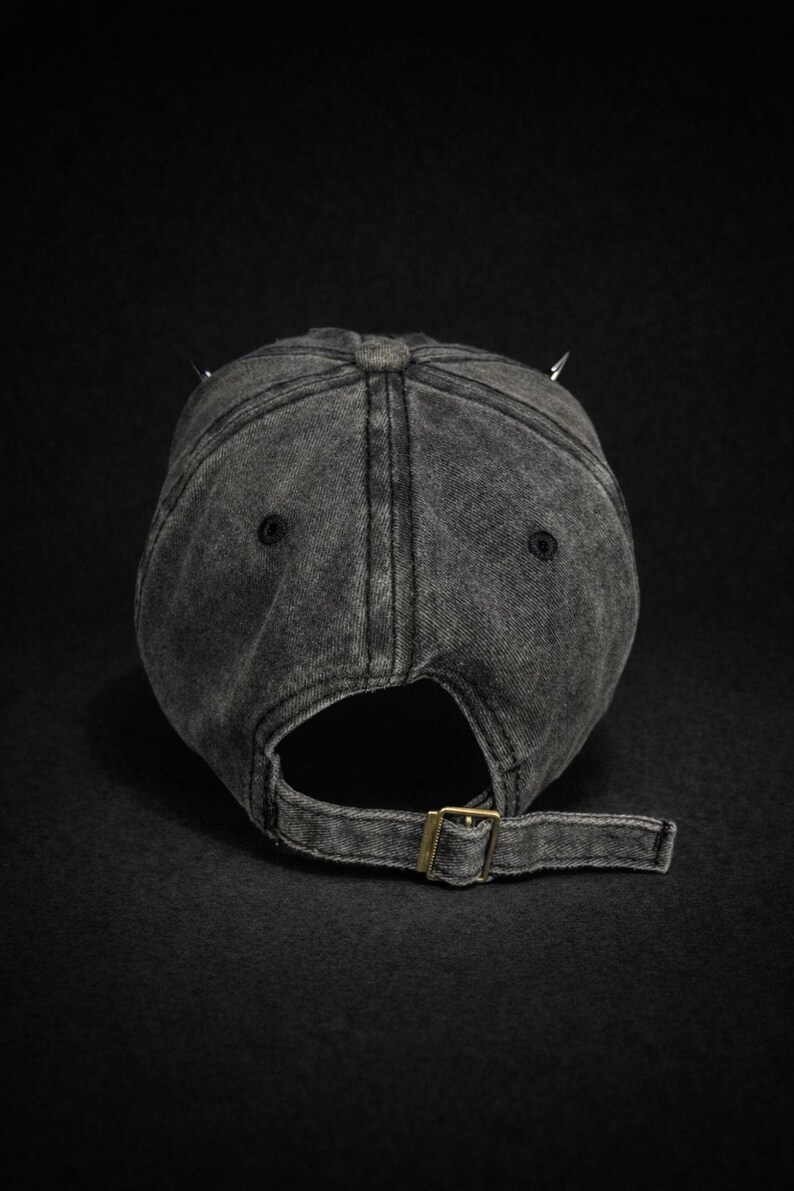 May include: A dark gray, distressed baseball cap with a curved brim and adjustable strap. The cap features two silver-colored spikes on top. The back of the cap has a metal buckle for adjustment.