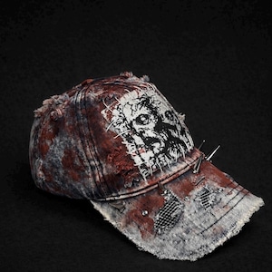 Pode incluir: Boné de beisebol de ganga desgastado com um gráfico de zumbi. O boné apresenta um esquema de cores vermelho e cinza desbotado, pontas de metal e um design rasgado e remendado. A palavra "ZURÜCK" é visível.
