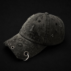 Puede incluir: Gorra de béisbol negra desgastada con detalles de anillos metálicos. La gorra presenta un aspecto desgastado y descolorido con rasgaduras y desgarros intencionales. La visera tiene varios anillos metálicos y la gorra está hecha de tela suave y texturizada.