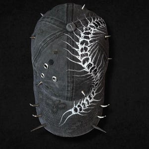 May include: A distressed charcoal gray baseball cap featuring a white graphic of a segmented centipede skeleton. The cap is adorned with multiple silver-toned metal spikes along the sides and brim, and includes several silver metal eyelet grommets.