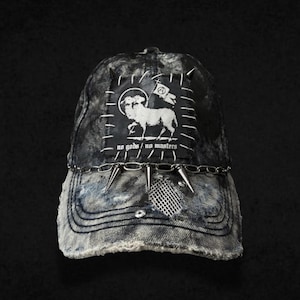 Può includere: Cappellino da baseball in denim effetto used, con una toppa raffigurante un agnello e la scritta "no gods / no masters". Il cappellino ha una catena e dettagli a punta sulla visiera. Il cappellino è blu scuro e grigio.