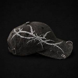 Könnte beinhalten: Schwarze Baseballkappe mit Used-Look und weißem Stacheldraht-Design. Die Kappe hat einen gebogenen Schirm mit silbernen Ringen und einen verstellbaren Riemen.