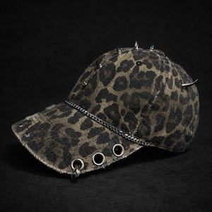 Op de afbeelding: Een baseballcap met luipaardprint, zilveren spikes en een kettingdetail. De pet heeft een versleten rand en metalen oogjes. De hoed is een mix van zwart en olijfgroen.