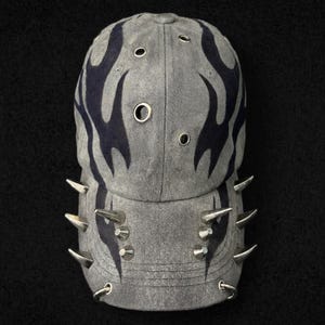 Peut inclure: Casquette de baseball grise avec un motif de flammes bleu foncé. La casquette est ornée de pointes argentées le long du bord et est fabriquée dans un matériau semblable au denim. La casquette comporte plusieurs trous d'aération.