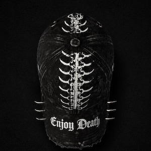 Könnte beinhalten: Eine schwarze Baseballkappe im Used-Look mit einer weißen Grafik einer menschlichen Wirbelsäule, die vertikal vom Schirm bis zum Kopf verläuft. Der Text "Enjoy Death" ist in weißer gotischer Schrift über dem Schirm gedruckt, ergänzt durch drei silberne Ringe.