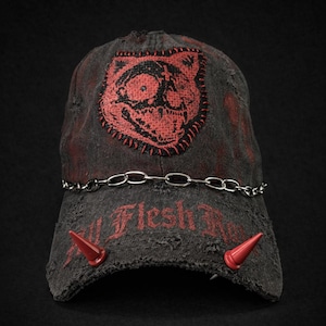 Könnte beinhalten: Verwaschene schwarze Baseballkappe mit einem roten, gestickten Katzen-Totenkopf-Aufnäher. Mit einer silbernen Kette über dem Schirm, roten Hörnern und dem Text "All Flesh Rots".