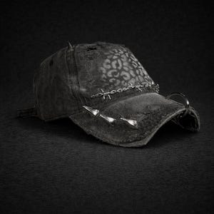 May include: A distressed black baseball cap with a leopard print pattern on the crown. The cap features silver-toned spikes and barbed wire detailing across the brim, with a metal ring attached to the side. The overall aesthetic is edgy and punk-inspired.