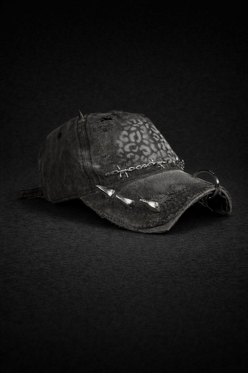 May include: A distressed black baseball cap with a leopard print pattern on the crown. The cap features silver-toned spikes and barbed wire detailing across the brim, with a metal ring attached to the side. The overall aesthetic is edgy and punk-inspired.