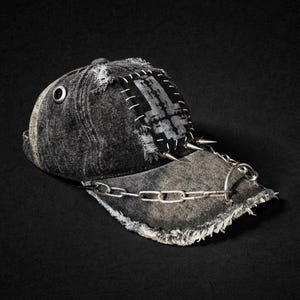 May include: Distressed black denim baseball cap with a silver chain and spikes. The cap features a patched design with white stitching and a metal eyelet. The brim is frayed, and the cap has a grunge aesthetic.