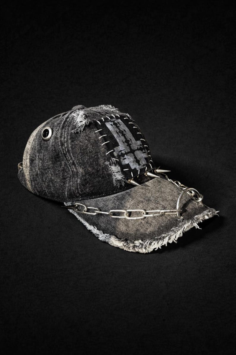 May include: Distressed black denim baseball cap with a silver chain and spikes. The cap features a patched design with white stitching and a metal eyelet. The brim is frayed, and the cap has a grunge aesthetic.