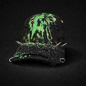 Könnte beinhalten: Schwarze Baseballkappe mit grünem Farbspritzer-Design und silbernen Stacheln. Die Kappe hat silberne Ösen und einen gebogenen Schirm. Der Look ist edgy und Punkrock.
