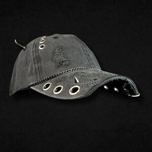 May include: A distressed charcoal grey baseball cap featuring silver-toned metal hardware, including multiple circular grommets, a thin chain across the brim, and pointed spikes. The fabric has a worn, textured appearance with frayed detailing.