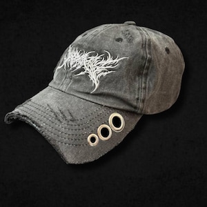 Op de afbeelding: Een verweerde antracietgrijze baseballpet met een wit geborduurd grafisch ontwerp op het voorpaneel. De gebogen klep bevat drie zilverkleurige metalen ringen op een rij, met gerafelde randen langs de klep en de bovenkant van de pet.