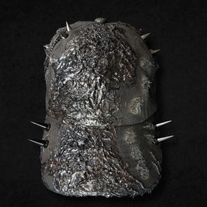 Op de afbeelding: Een donkergrijze baseballpet met een zwaar verweerd, getextureerd oppervlak met metalen, gesmolten details op de voorpanelen. De pet is versierd met meerdere scherpe, zilverkleurige metalen spikes langs de zijkanten en de bovenkant.