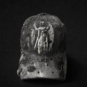 Może przedstawiać: Zniszczona czarna czapka bejsbolowa z białą grafiką anioła. Czapka posiada srebrny łańcuszek, metalowe ćwieki i zakrzywiony daszek. Kapelusz jest umieszczony na czarnym tle.