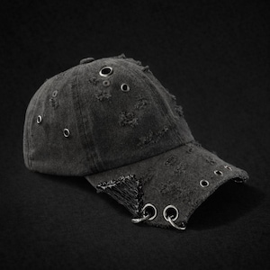 Puede incluir: Gorra de béisbol negra desgastada con un diseño rasgado. La gorra presenta ojales metálicos y un borde deshilachado, lo que le da una estética robusta y atrevida. El sombrero está sobre un fondo oscuro.