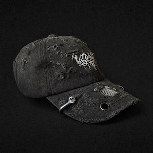 Pode incluir: Boné de beisebol preto desgastado com aba curva. O boné apresenta um design rasgado, ilhós de metal e um anel de metal prateado na aba. Texto branco bordado na frente. O boné está sobre um fundo preto.