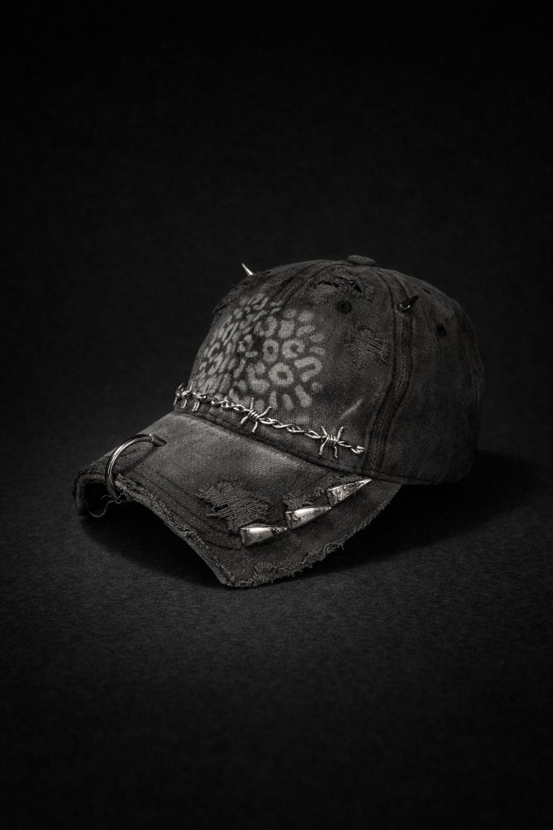 May include: Black baseball cap with distressed details, metal spikes, and barbed wire accents. The cap features a faded leopard print design on the crown and a metal ring on the brim. A stylish and edgy accessory.