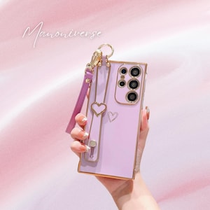 May include: A lavender phone case with a gold-colored frame and heart accents. The case has a matching lavender wrist strap and a gold-colored ring holder. The text "Manoniverse" is written in cursive on the top left.
