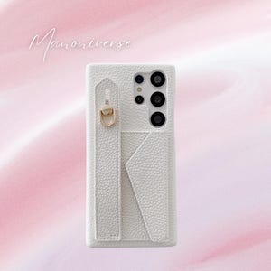 May include: White textured phone case with a strap and a gold-colored ring. The case has a cardholder and a camera cutout. The text "Manoniverse" is written in cursive on the top left corner.