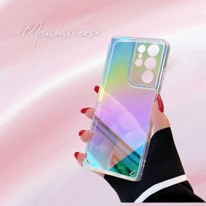 Op de afbeelding: Een doorzichtige telefoonhoes met een iriserend regenboogeffect. De hoes wordt vastgehouden in een hand met rode nagellak. De tekst "Manoniverse" staat cursief in de linkerbovenhoek. De hoes heeft een transparante rand en een camera-uitsparing.