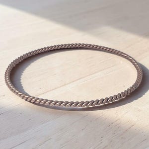 May include: A silver-colored, twisted metal bangle bracelet. The bracelet has a rope-like design and is circular in shape. The bracelet is resting on a light-colored wooden surface.
