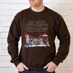 Pode incluir: Um moletom marrom com o logotipo de Star Wars adornado com luzes coloridas. O design apresenta personagens de Star Wars com gorros de Papai Noel, como R2-D2 e Yoda. O tema &eacute; festivo e natalino.