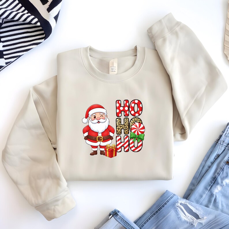 Op de afbeelding: Een cr&egrave;mekleurige sweatshirt met een kerstontwerp. Het heeft een knipogende kerstman, een rode geschenkdoos en de woorden "HO HO HO" in verschillende patronen. Het ontwerp bevat stippen, luipaardprint en zuurstokstrepen, met een groene strik.