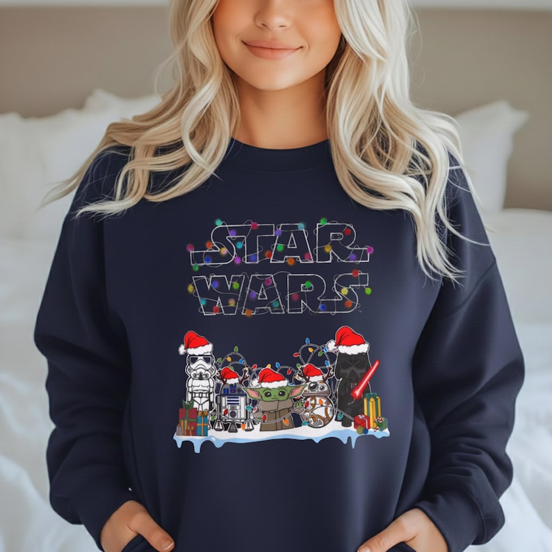 Pode incluir: Camisola azul marinho com um design festivo de Star Wars. O texto "STAR WARS" &eacute; delineado com luzes de Natal coloridas. O design inclui personagens como Stormtrooper, R2-D2, Grogu e Darth Vader, todos com gorros de Pai Natal.
