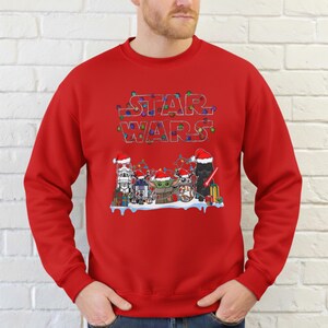 Pode incluir: Uma camisola vermelha com um design de Star Wars. A palavra "STAR WARS" est&aacute; escrita no topo em uma fonte festiva com luzes de Natal. Abaixo, personagens de Star Wars com chap&eacute;us de Papai Noel.