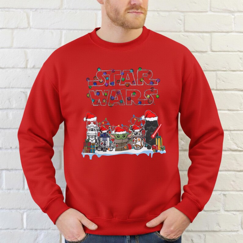 Pode incluir: Uma camisola vermelha com um design de Star Wars. A palavra "STAR WARS" est&aacute; escrita no topo em uma fonte festiva com luzes de Natal. Abaixo, personagens de Star Wars com chap&eacute;us de Papai Noel.