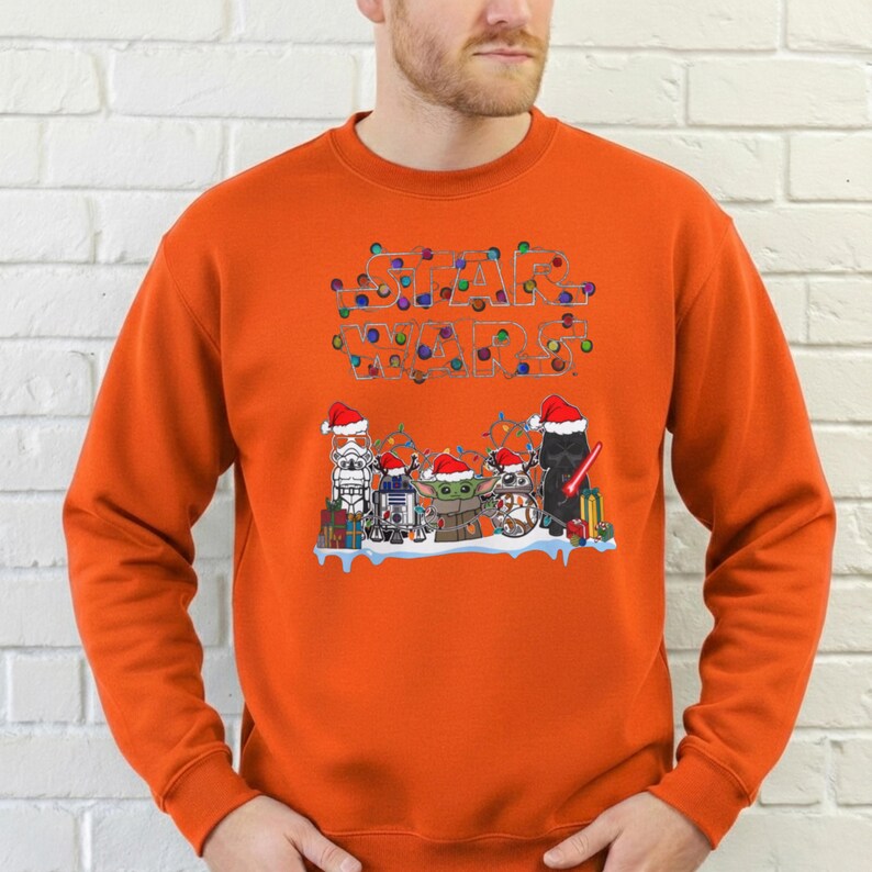 Pode incluir: Camisola laranja com um design de Star Wars. O design apresenta personagens com chap&eacute;us de Pai Natal, com as palavras "STAR WARS" escritas com luzes de Natal. Os personagens incluem Stormtrooper, R2-D2, Yoda e Darth Vader.