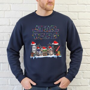 Pode incluir: Camisola azul marinho com o log&oacute;tipo Star Wars adornado com luzes coloridas. O design apresenta personagens como Stormtrooper, R2-D2, Grogu e Darth Vader, todos com gorros de Pai Natal. H&aacute; presentes na parte inferior da imagem.