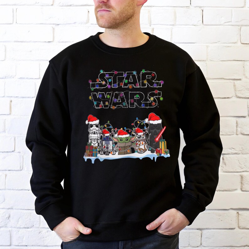 Pode incluir: Camisola preta com o log&oacute;tipo Star Wars decorado com luzes coloridas. Abaixo do log&oacute;tipo, personagens de Star Wars com gorros de Pai Natal, incluindo Stormtrooper, R2-D2, Grogu e outros. Presentes tamb&eacute;m est&atilde;o inclu&iacute;dos.