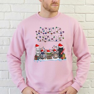 Pode incluir: Camisola rosa claro com um design de Star Wars. O design apresenta luzes festivas e personagens com chap&eacute;us de Pai Natal, incluindo R2-D2, Grogu e Darth Vader, criando um tema de f&eacute;rias.