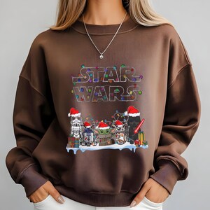 Pode incluir: Um moletom marrom com o logotipo de Star Wars decorado com luzes de Natal. O design inclui personagens de Star Wars com chap&eacute;us de Papai Noel: Stormtrooper, R2-D2, Grogu e Darth Vader.
