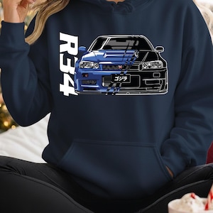 May include: Navy blue hoodie featuring a split graphic of a blue and black sports car. The white text "R34" is vertically aligned on the left side. The car graphic includes Japanese text. A comfortable, casual garment.