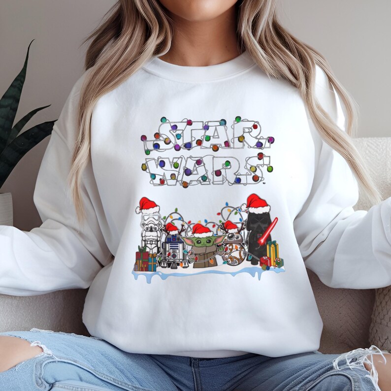Pode incluir: Camisola branca com um design de Natal de Star Wars. O texto "STAR WARS" &eacute; iluminado por luzes coloridas. Abaixo, v&aacute;rios personagens de Star Wars usam chap&eacute;us de Papai Noel e est&atilde;o rodeados de presentes.