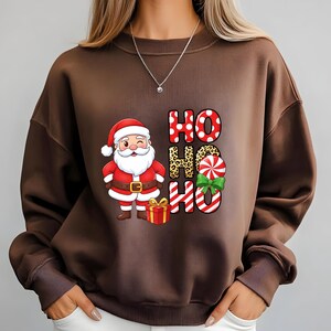 Op de afbeelding: Een bruine sweatshirt met een kerstman-ontwerp en de tekst "HO HO HO". De Santa-illustratie is in een rood pak met een witte baard en hoed. De letters "HO" zijn in rode stippen, luipaardprint en zuurstokstrepen.