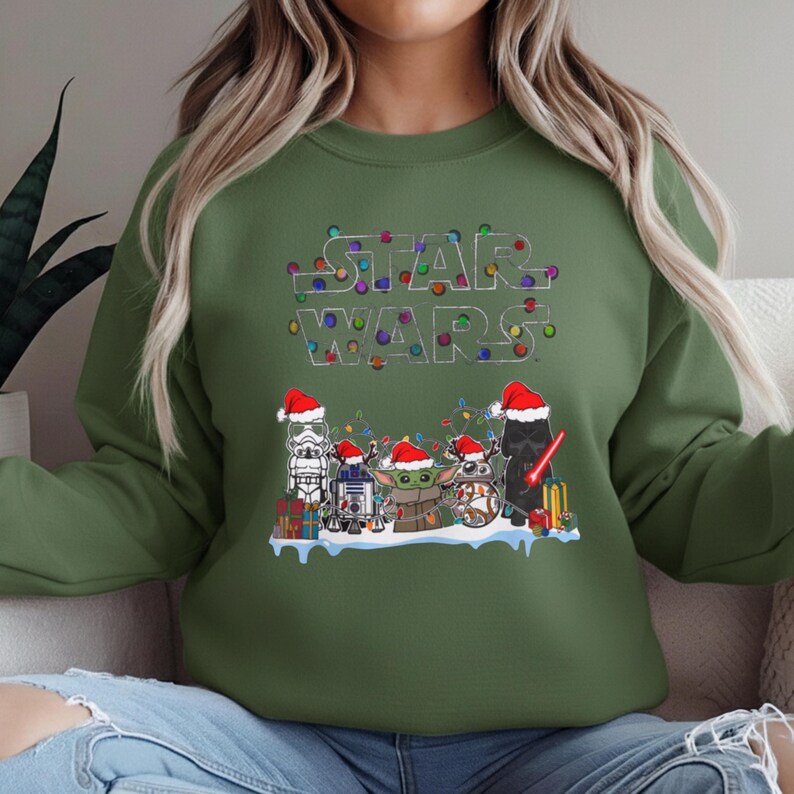 Pode incluir: Camisola verde com o log&oacute;tipo Star Wars decorado com luzes de Natal coloridas. Abaixo do log&oacute;tipo, personagens de Star Wars com gorros de Pai Natal: um Stormtrooper, R2-D2, Grogu e Darth Vader. Um design festivo para as festas.