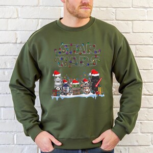Pode incluir: Camisola verde-oliva com um design de Natal Star Wars. O gr&aacute;fico inclui o logotipo Star Wars com luzes coloridas e personagens como Stormtrooper, R2-D2, Grogu e Darth Vader, todos usando chap&eacute;us de Papai Noel.