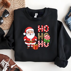 Op de afbeelding: Zwarte sweatshirt met een Santa Claus-afbeelding en de woorden "HO HO HO". De Santa-illustratie heeft een rood pak, een witte baard en een geschenkdoos. De "HO" letters zijn voorzien van stippen en luipaardprint.