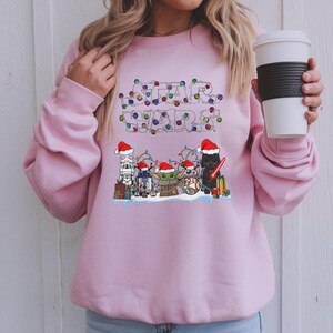 Pode incluir: Um moletom rosa claro com a inscri&ccedil;&atilde;o "STAR WARS" em um design de luzes festivas e coloridas. Abaixo do texto, personagens de desenhos animados da franquia Star Wars com gorros de Papai Noel, incluindo Darth Vader e Baby Yoda.