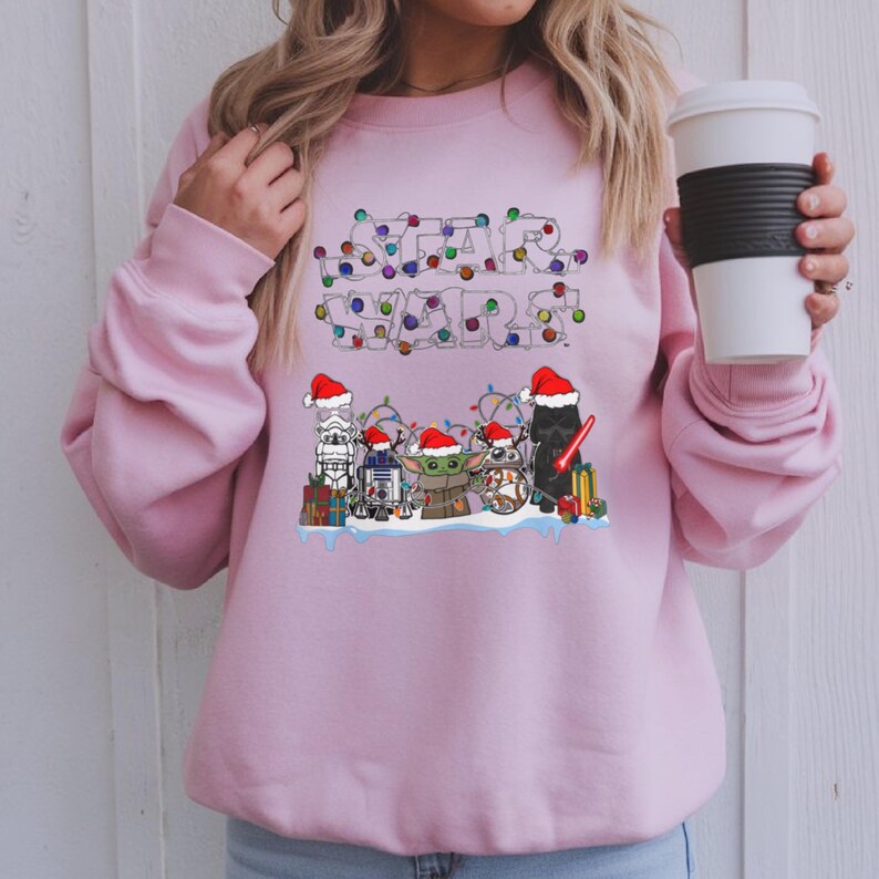 Pode incluir: Um moletom rosa claro com a inscri&ccedil;&atilde;o "STAR WARS" em um design de luzes festivas e coloridas. Abaixo do texto, personagens de desenhos animados da franquia Star Wars com gorros de Papai Noel, incluindo Darth Vader e Baby Yoda.