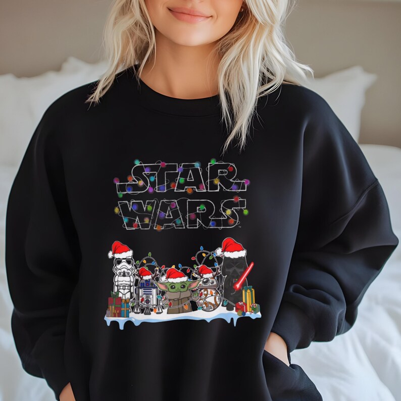 Pode incluir: Camisola preta com o log&oacute;tipo Star Wars decorado com luzes de Natal. O design inclui personagens de Star Wars com gorros de Pai Natal: um Stormtrooper, R2-D2, Grogu e Darth Vader.