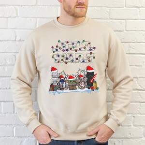 Pode incluir: Camisola bege com um design de Star Wars. A palavra "STAR WARS" &eacute; formada com luzes de Natal coloridas. Personagens como Stormtrooper, R2-D2, Grogu e Darth Vader usam chap&eacute;us de Papai Noel.