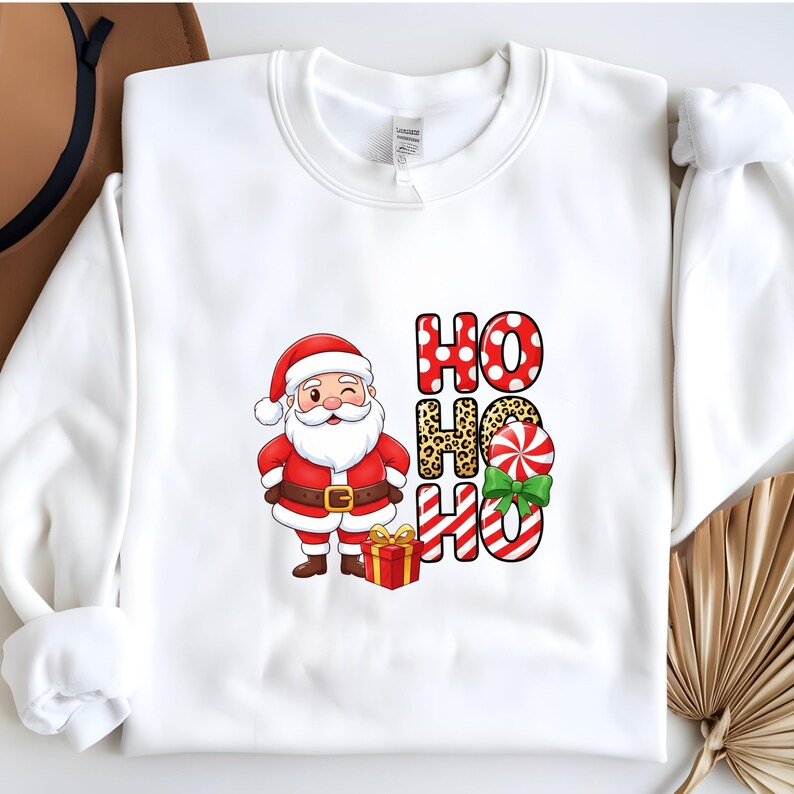 Op de afbeelding: Witte sweatshirt met een cartoon kerstman die knipoogt, een rood pak en hoed draagt en een cadeau vasthoudt. De woorden "HO HO HO" zijn gedrukt in rode stippen, luipaardprint en zuurstokstrepen.