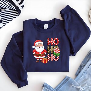 Op de afbeelding: Marineblauw sweatshirt met een kerstman-afbeelding en de woorden "HO HO HO". De kerstman-illustratie bevat een rood pak, een witte baard en een geschenkdoos. De tekst "HO" is in rode stippen, luipaardprint en zuurstokstrepen.