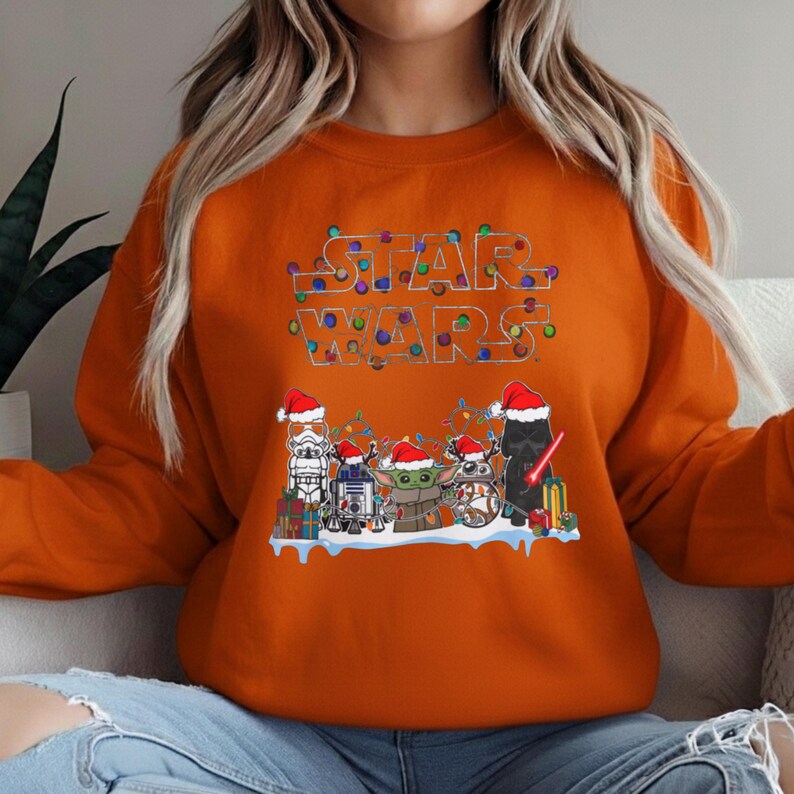 Pode incluir: Camisola laranja com um design festivo de Star Wars. O texto "STAR WARS" &eacute; iluminado com luzes de Natal coloridas. Abaixo, v&aacute;rios personagens de Star Wars usam chap&eacute;us de Papai Noel e est&atilde;o rodeados de presentes.